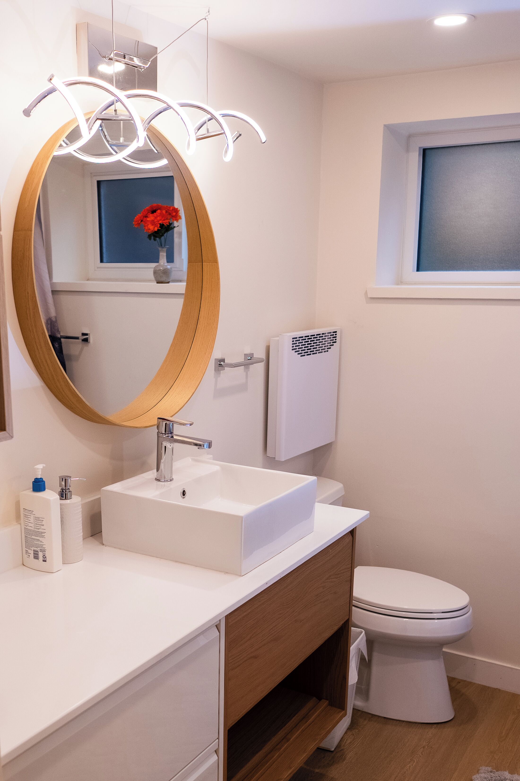 A modern bathroom with a white sink, a wooden vanity, a round mirror, a white toilet, and a window with a view.