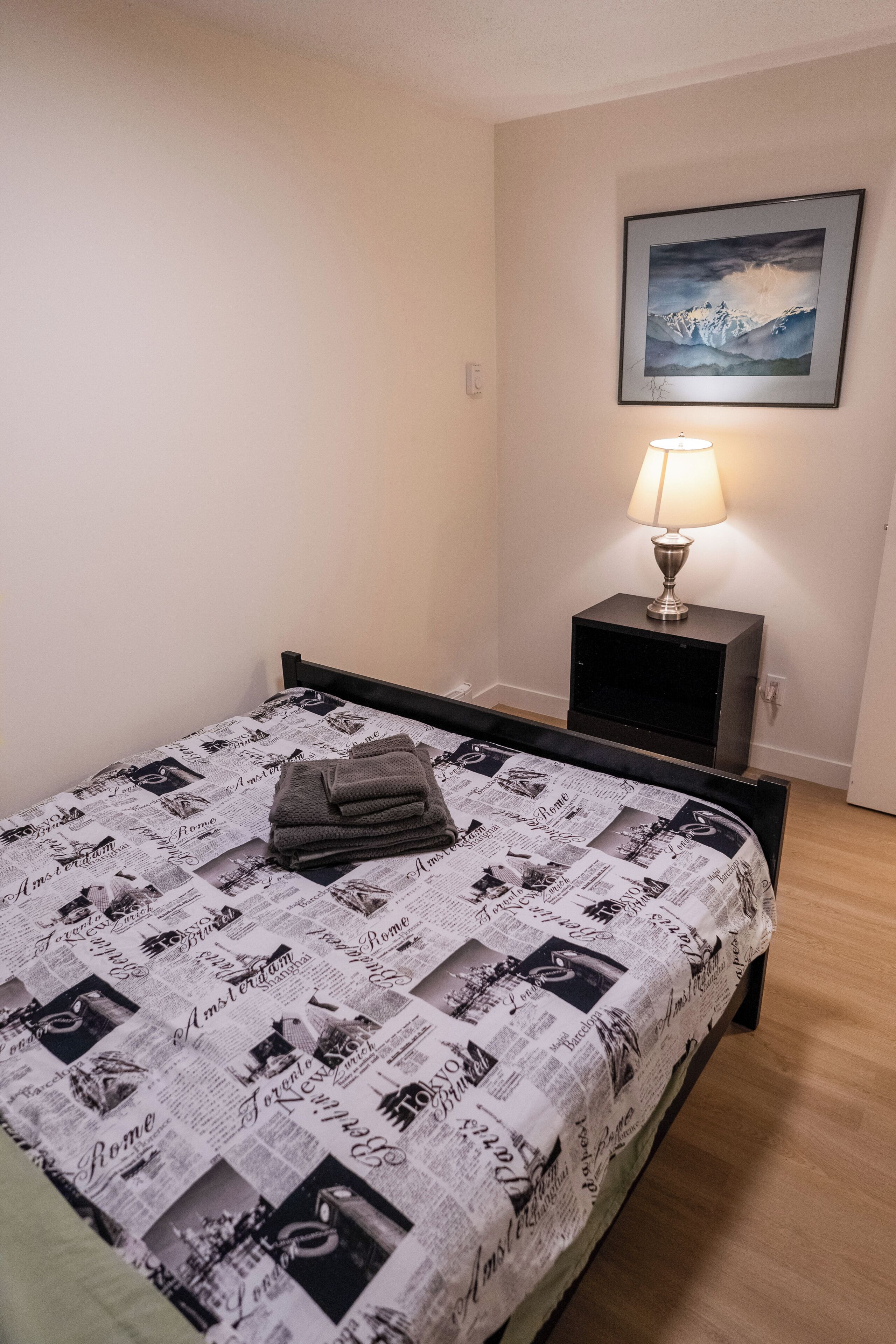 Ein ordentlich bezogenes Bett mit zeitungsmotivbedeckter Decke, eine Nachttischlampe und ein gerahmtes Bild an der Wand.