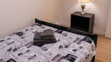 Ein ordentlich bezogenes Bett mit zeitungsmotivbedeckter Decke, eine Nachttischlampe und ein gerahmtes Bild an der Wand.