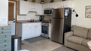 Une cuisine compacte avec des meubles blancs, des appareils en acier inoxydable, un four à micro-ondes, un réfrigérateur, une cuisinière et un évier. On y trouve également une petite poubelle et un tapis sur le sol.