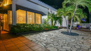 A modern building with large windows, a stone-paved courtyard, and a well-maintained garden with palm trees and flowering plants. - Luxury pool villa totally private backyard - 5 minutes to the beach! (Noord)
