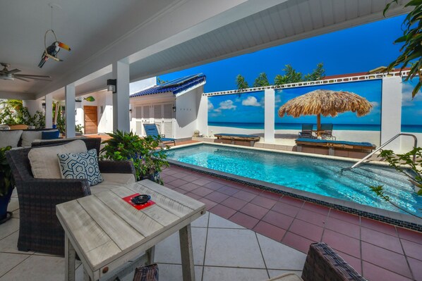Zona de piscina con vista al océano, una sombrilla de paja y sillones de descanso.