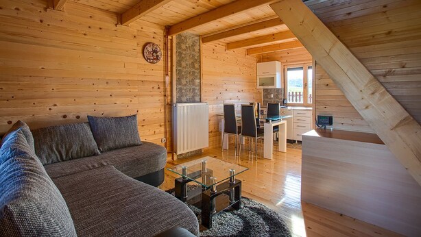 Ein gemütliches Blockhaus-Interieur mit Wohnbereich, der eine Couch und einen Glastisch, eine Wand mit Steinakzenten, einen Essbereich mit Stühlen und einem Tisch sowie eine Küche mit einer Mikrowelle zeigt.
