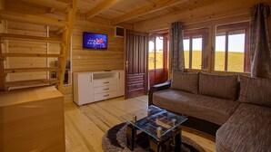 Ein gemütliches Blockhaus-Interieur mit Holzboden, einer Couch, einem Glastisch, einem Fernseher und einem großen Fenster mit Vorhängen.