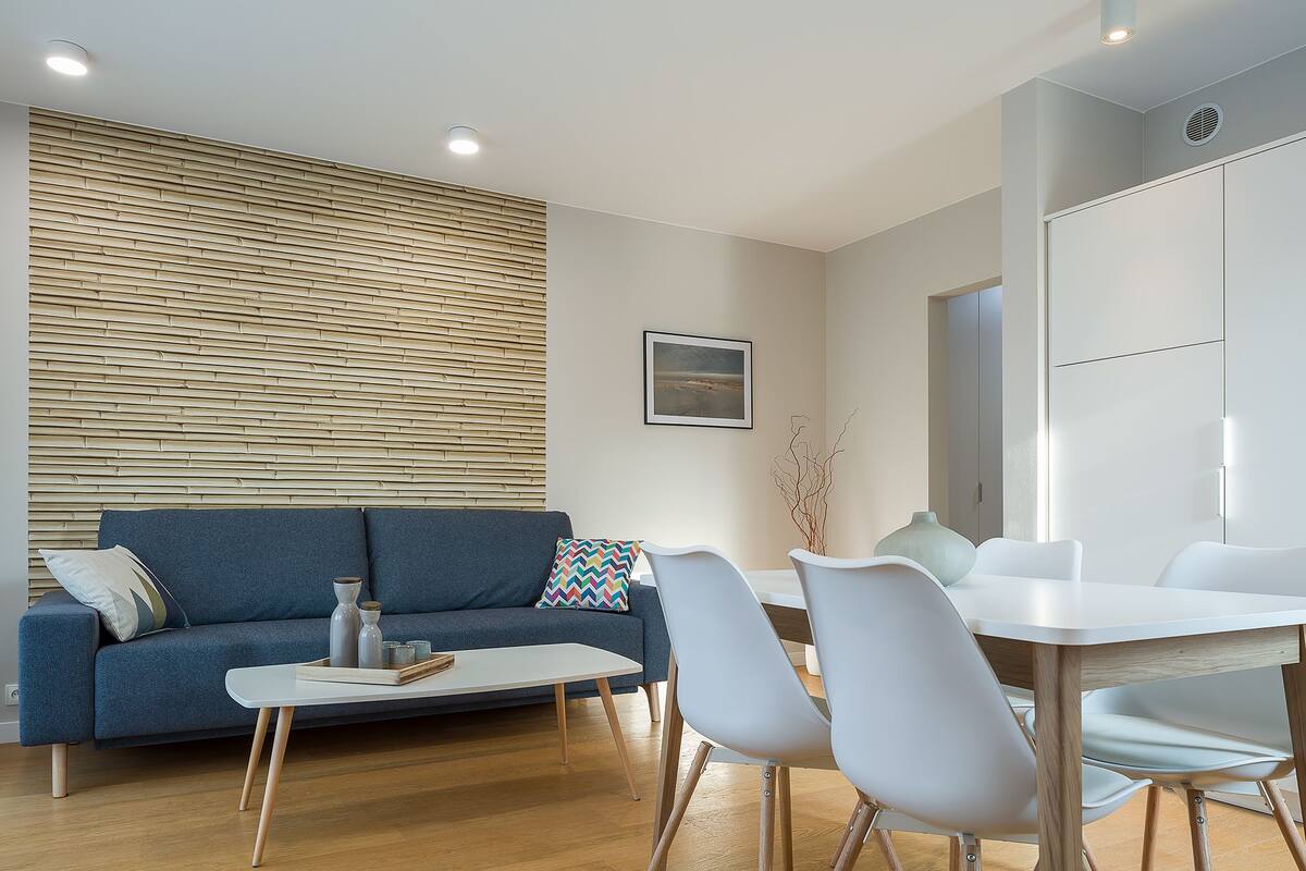 A modern living room with a blue sofa, a white coffee table, and a dining area with a white table and grey chairs.