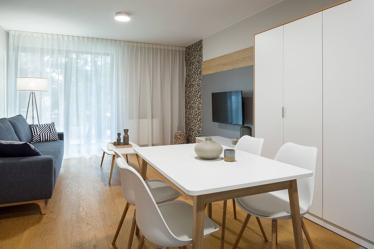 A modern living room with a white dining table, white chairs, a grey sofa, and a TV mounted on the wall.