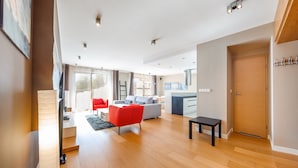 A modern living room with a grey sofa, red armchair, and a small black table. There is a kitchen area with a stove and cabinets visible. - Apartamenty Sun&Snow Wczasowa Południowe (Ustka)