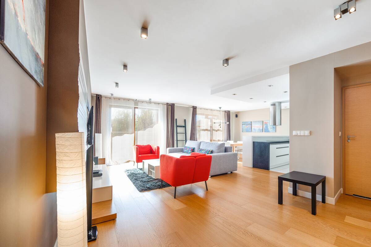 A modern living room with a grey sofa, red armchair, and a small black table. There is a kitchen area with a stove and cabinets visible.