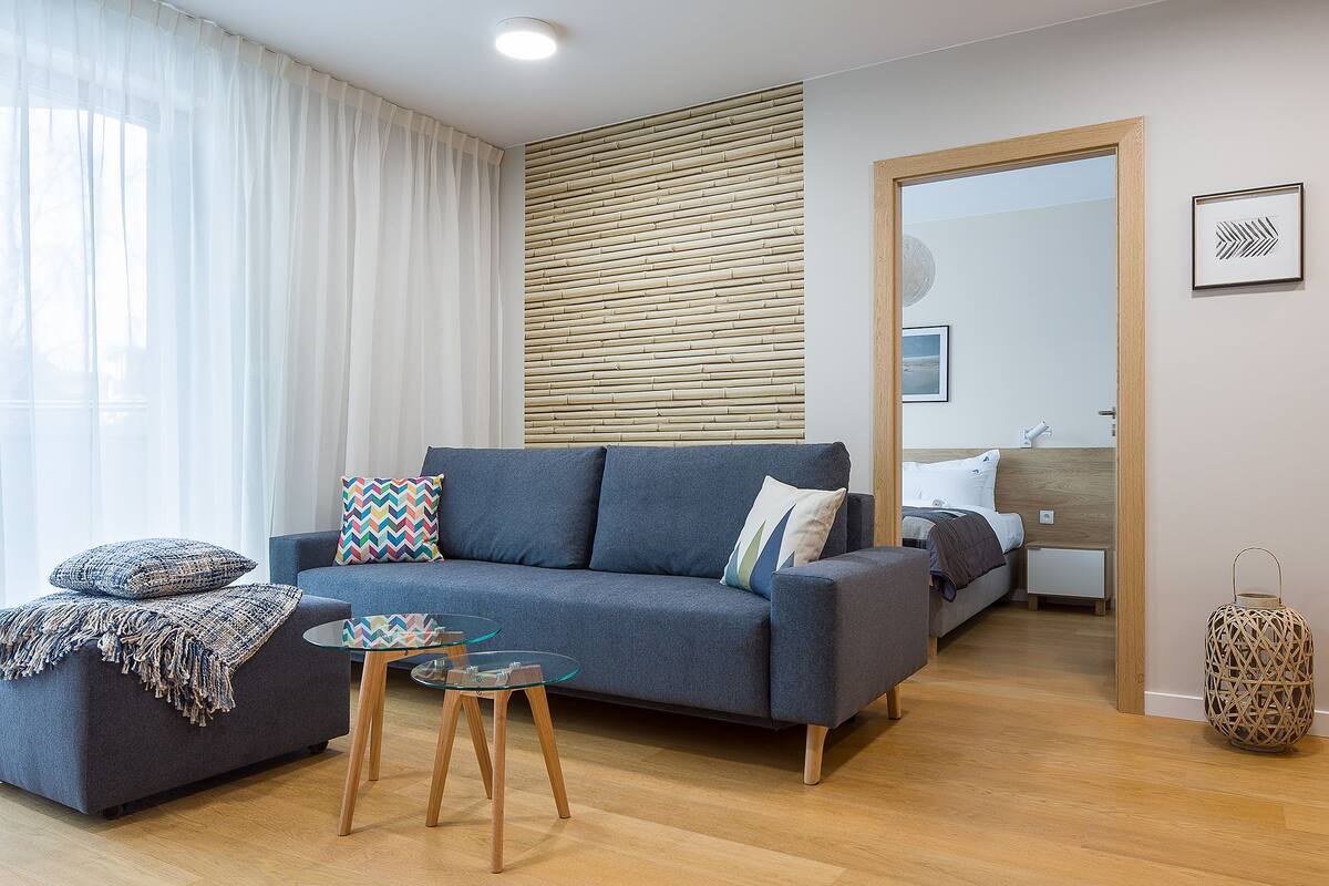 A modern living room with a grey sofa, two glass tables with wooden legs, a wooden wall panel, a bed with a wooden headboard, and a mirror on the wall.