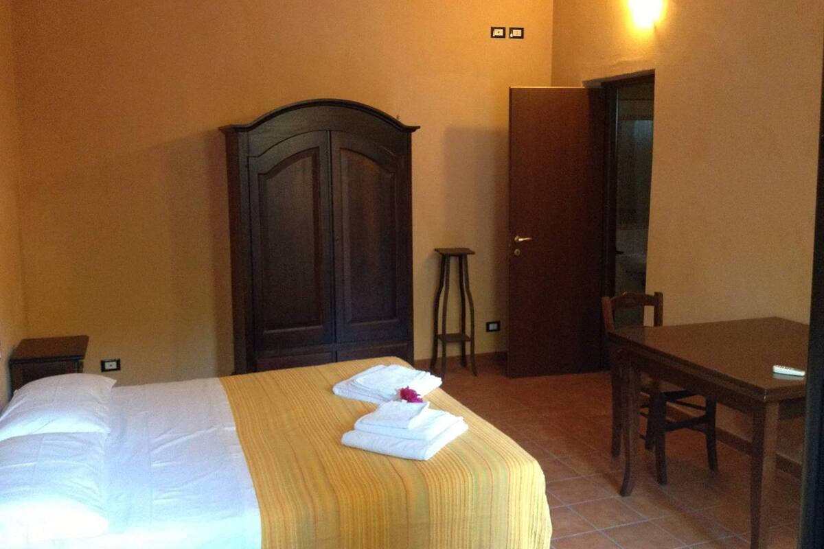 A hotel room with a bed, a wooden wardrobe, a small stool, a table, and a chair.