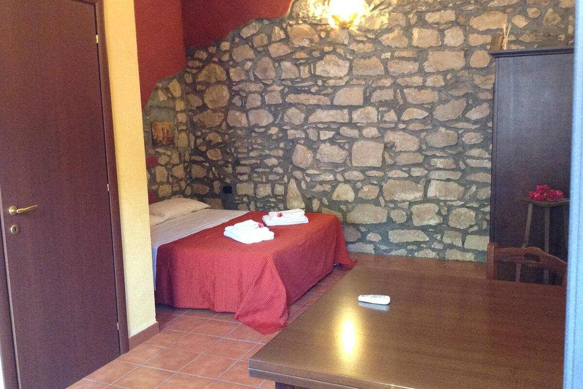A bedroom with a stone wall, a bed with red bedding, a wooden table, and a wardrobe.