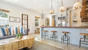 Un salon spacieux avec un canapé, une table basse et des tabourets de bar. On peut apercevoir une cuisine avec un comptoir-bar et des luminaires suspendus.