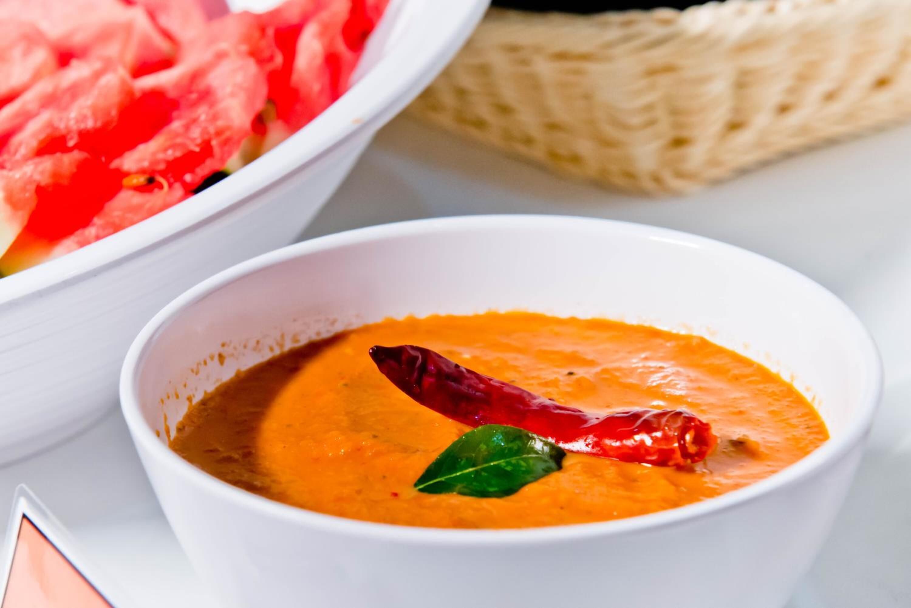 A bowl of orange soup with a red chili and a green leaf garnish, a basket of sliced watermelon, and a white bowl in the background.