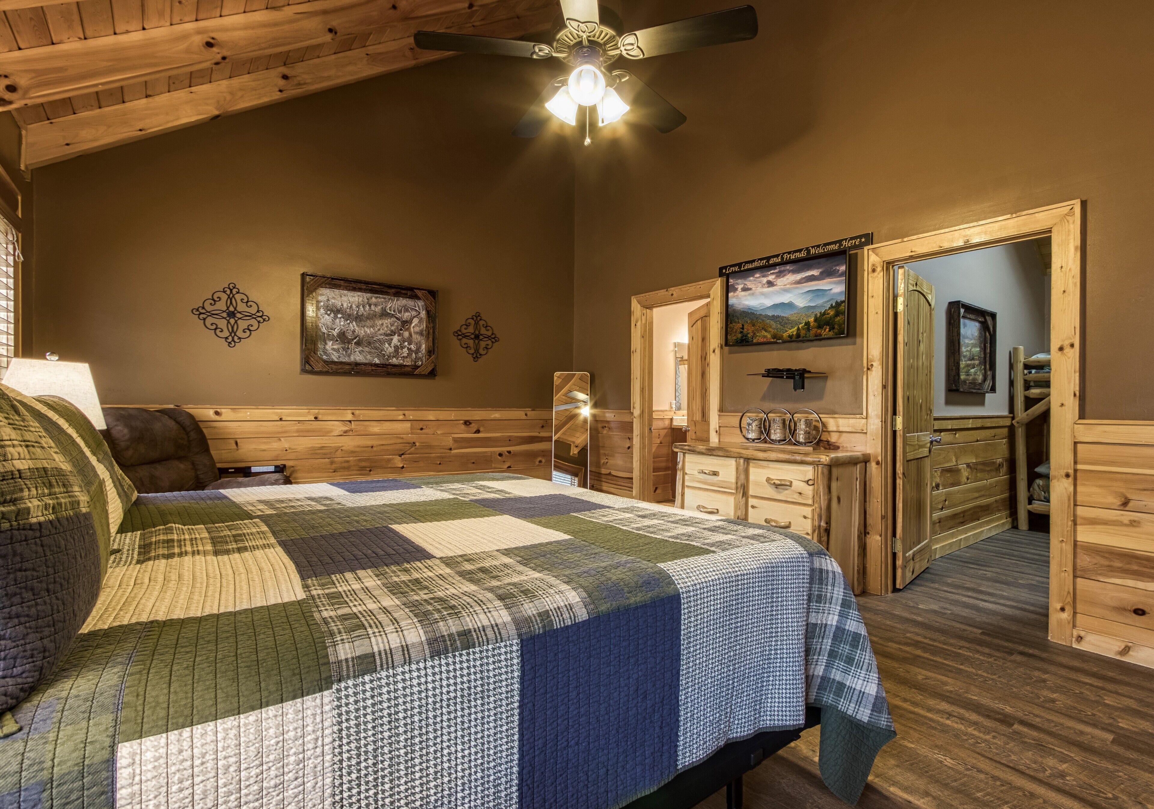 A bedroom with a bed, wooden ceiling, a ceiling fan, a nightstand, a dresser, a mirror, and a door leading to another room.