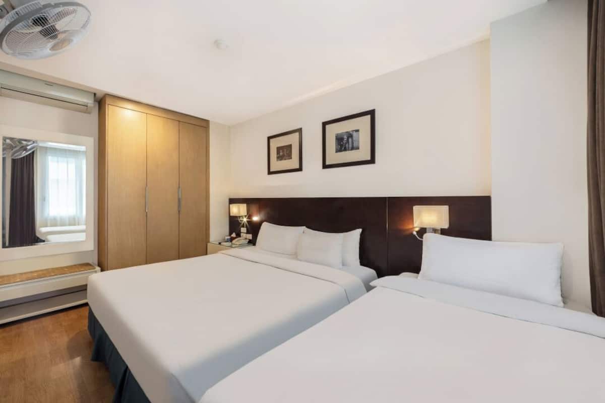 A hotel room with two beds, a wooden wardrobe, a ceiling fan, and two framed pictures on the wall.
