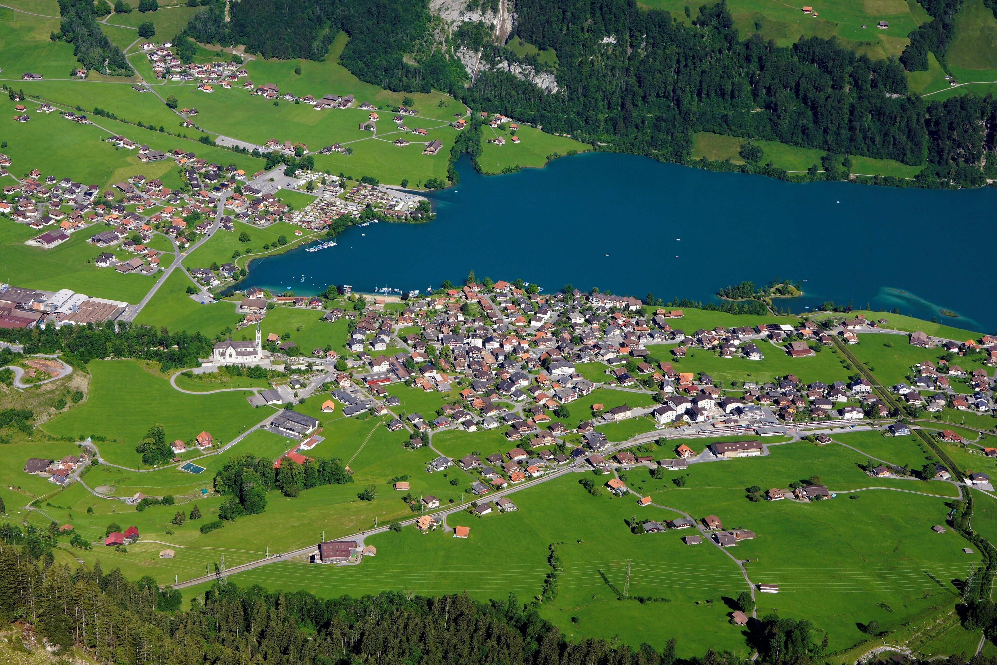 Vista aerea di una piccola cittadina con un lago, circondata da campi verdi e zone residenziali.