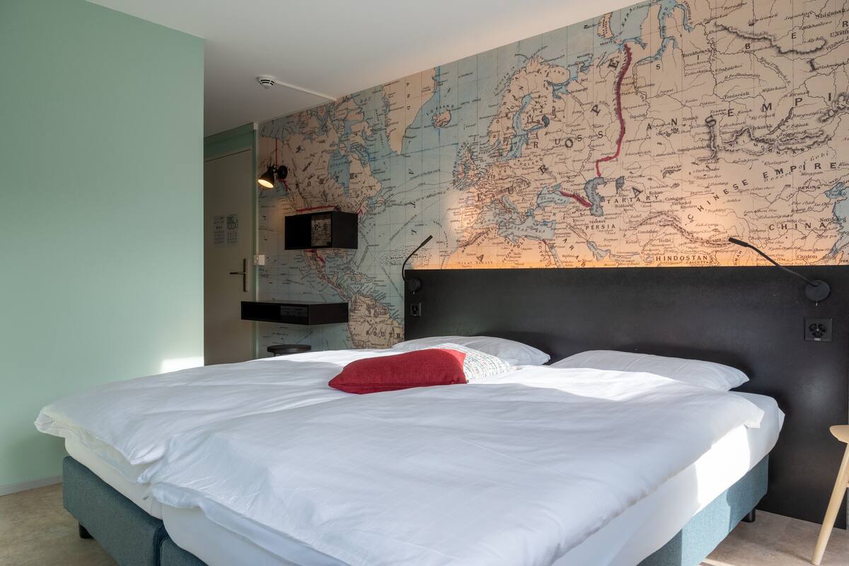 Un dormitorio con un gran mapa en la pared, una cama con ropa blanca y almohada roja, y una mesita de noche pequeña.
