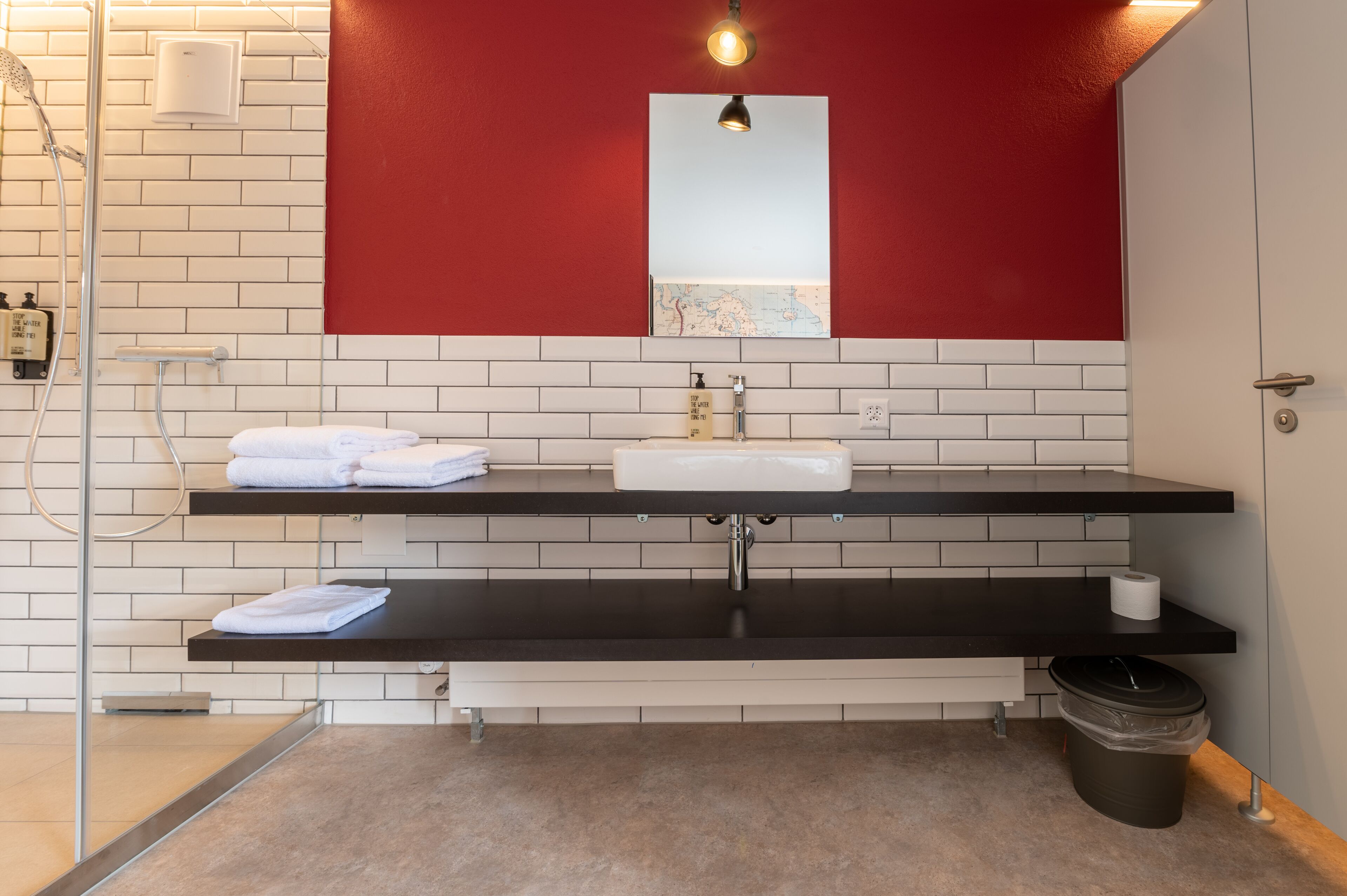 Une salle de bain moderne avec un mur recouvert de carreaux blancs, un plan de travail noir, un lavabo blanc et un mur d’accent rouge.