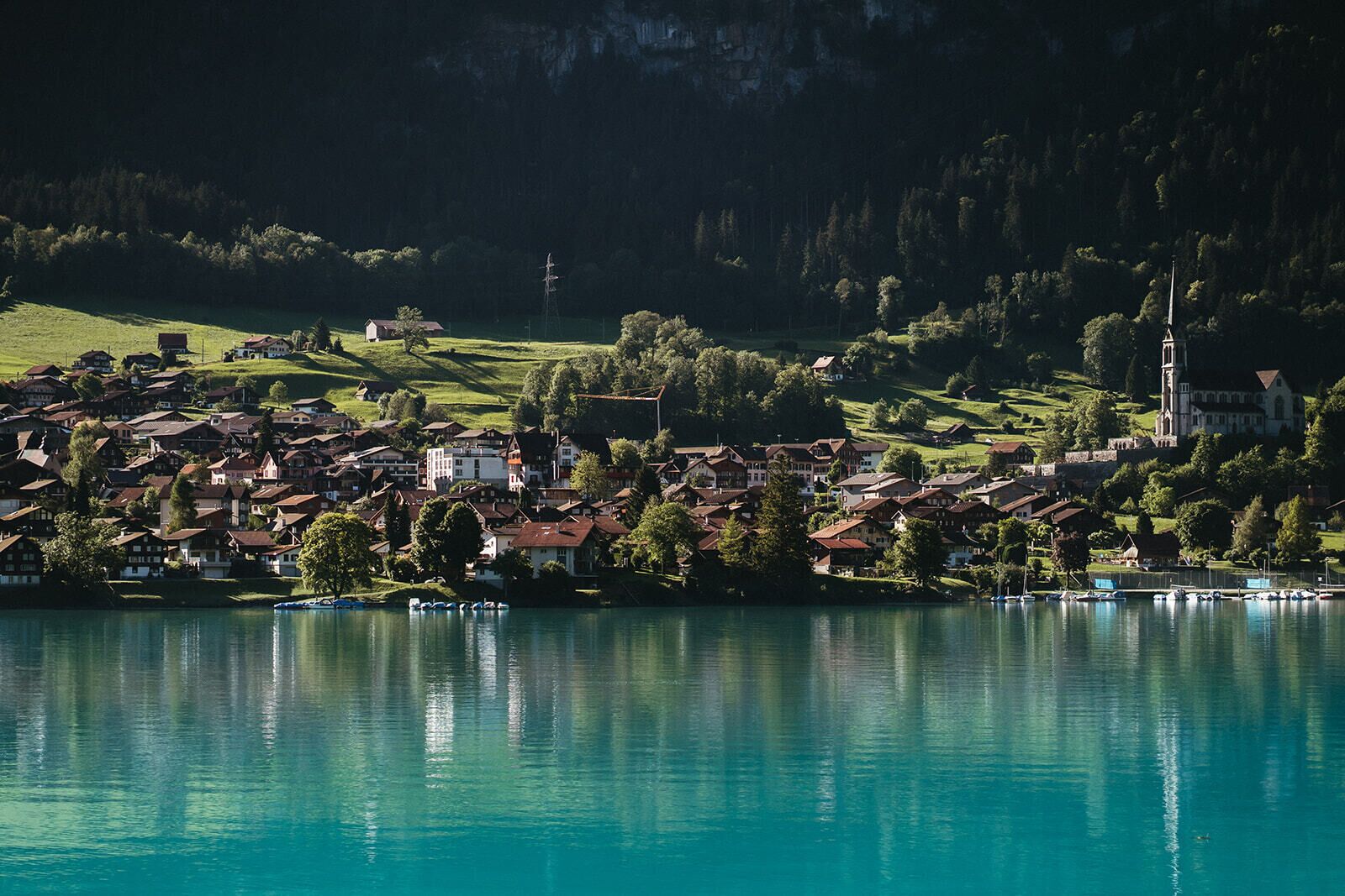 Un pueblo junto al lago con edificios a lo largo de la orilla, una iglesia con campanario y barcos amarrados en el muelle.