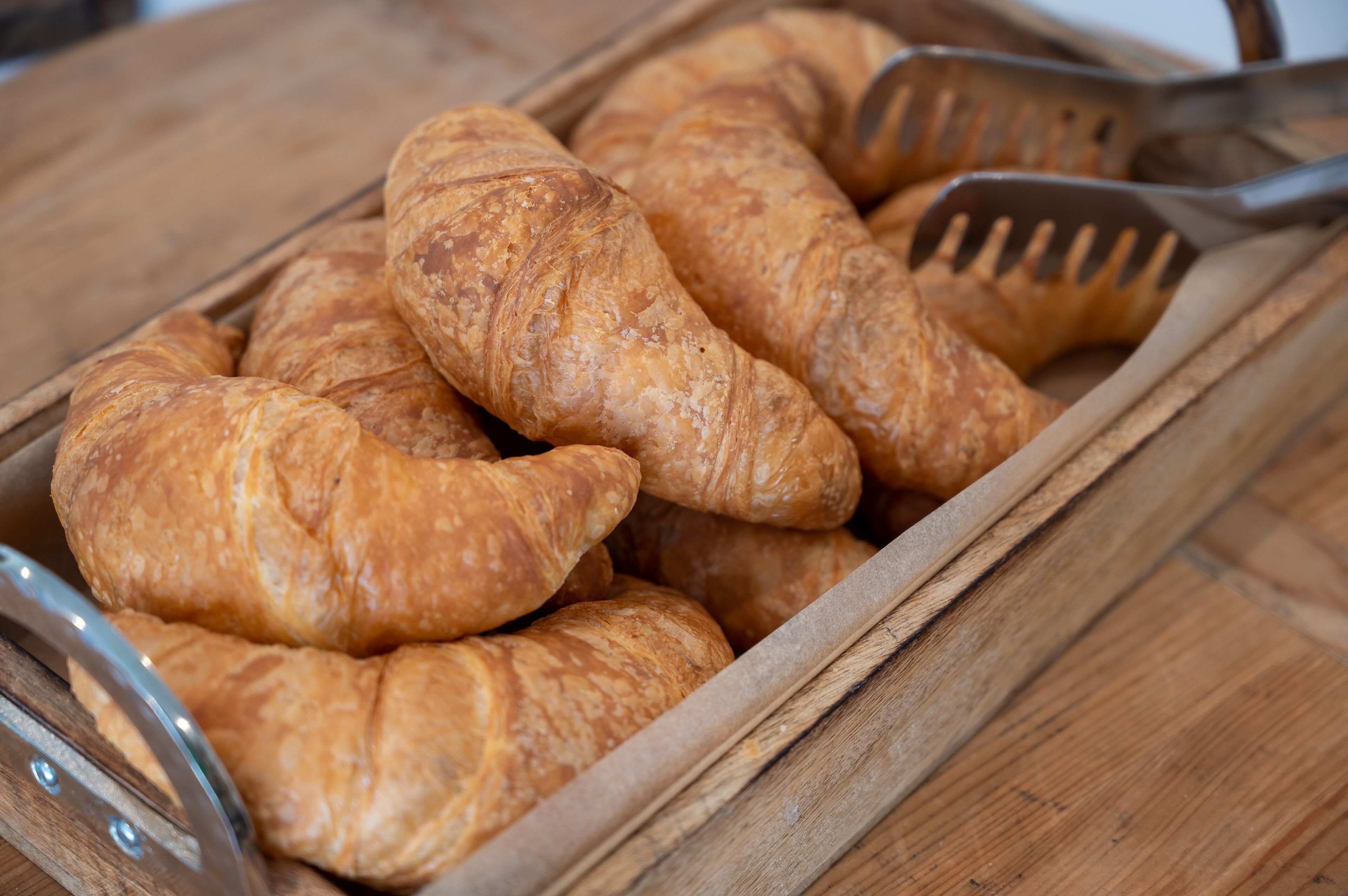 Uma bandeja de madeira com vários croissants dourados e um par de pinças de metal.
