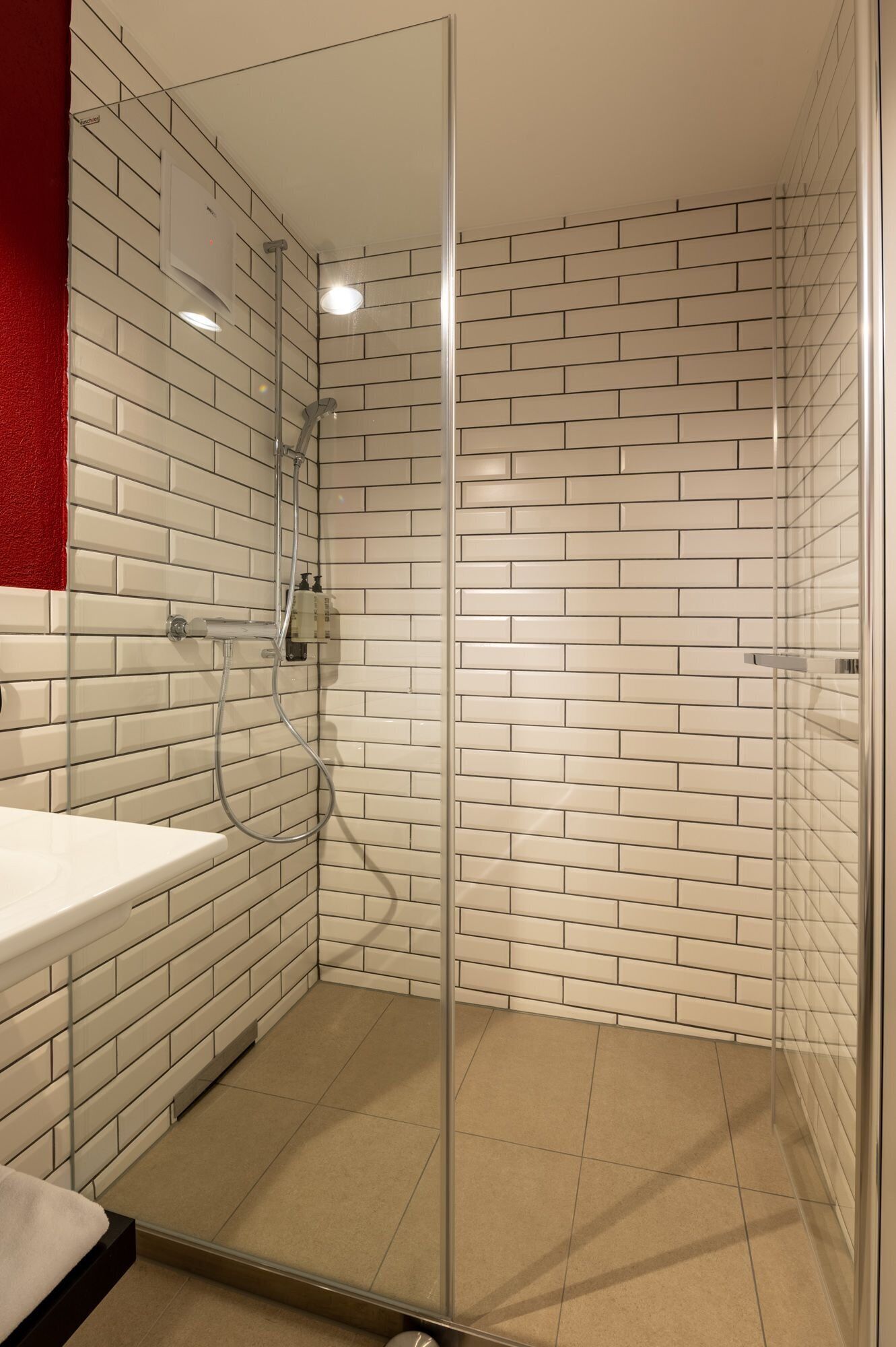 Une salle de bain moderne avec une douche à l’italienne en verre, des carreaux de céramique blancs rectangulaires et un mur d’accent rouge.