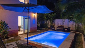 Una piscina con un elemento acuático iluminado en azul, rodeada de mobiliario exterior y un patio cubierto.