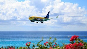 Un avion aux couleurs bleu et jaune survole une zone côtière, avec une eau d’un bleu limpide et une plage au premier plan.