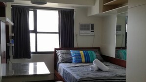 Una moderna camera d'albergo con un letto, una scrivania e una sedia. Si vede una finestra con tende, un condizionatore d'aria e una ventola a soffitto.