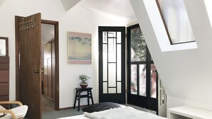 Une chambre à coucher avec un lit, une porte en bois, une petite table avec une plante et une fenêtre donnant sur l’extérieur.