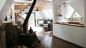 Un espace de vie de style loft avec un escalier en bois, une cheminée en pierre et un coin cuisine doté d’une fenêtre donnant sur un balcon.