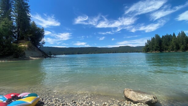 Un lac aux eaux limpides, bordé d’une rive rocheuse et entouré d’une forêt dense et de collines.