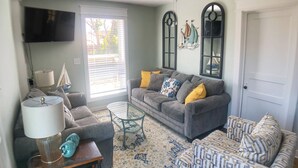 Ein Wohnzimmer mit grauem Sofa, einem Glastisch, einem Ottomanen mit Muster und einer Lampe.