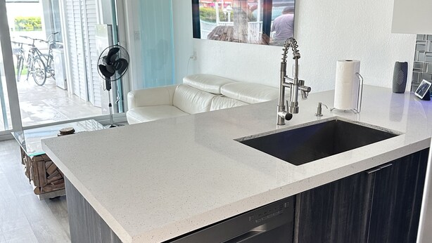 A modern kitchen with a stainless steel dishwasher, a large island with a sink, and a view of the outdoors through a glass door. - Dream Vacation Home Key Colony Beach Marathon Now with private pool! (Marathon)