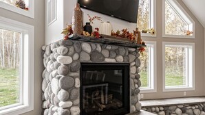 A stone fireplace with a flat-screen TV above it, large windows, and a stone-tiled floor.