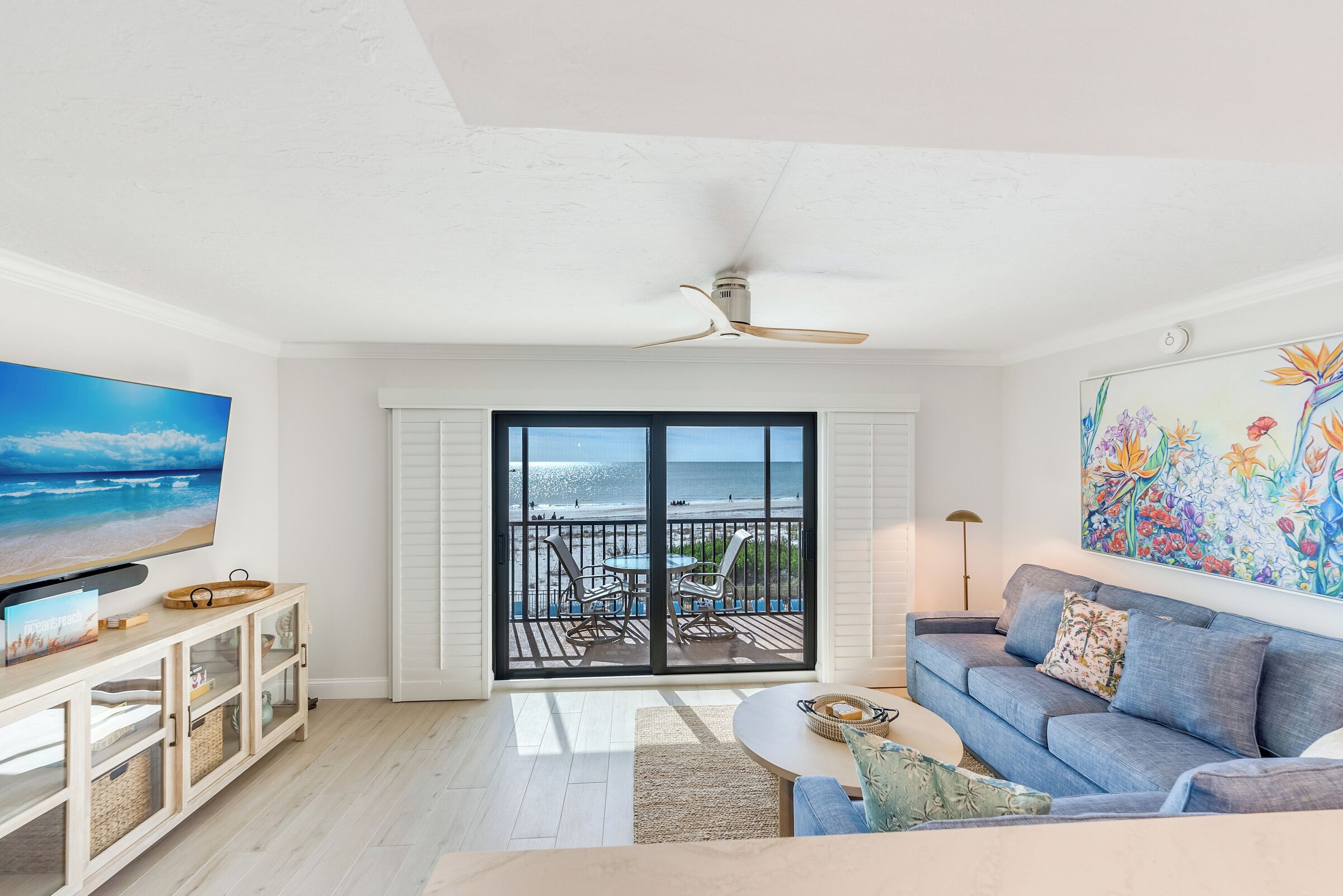 Ein Wohnzimmer mit großem Fernseher, einer Couch, einem Couchtisch und einem Balkon mit Blick auf den Strand.