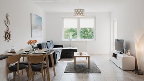 Een moderne woonkamer met een eethoek, een bank, een televisie en een raam met uitzicht op groen.