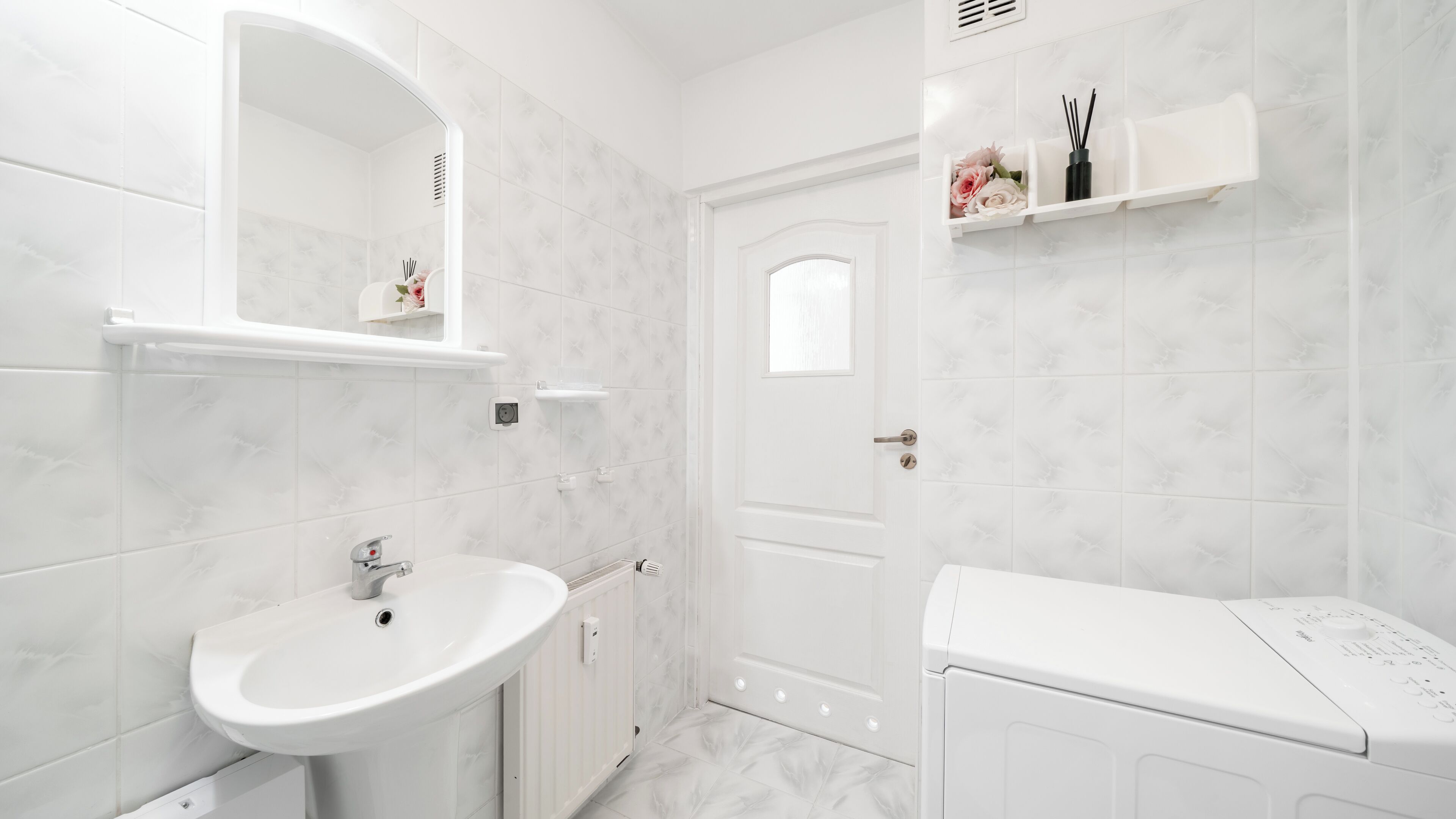 Ein Badezimmer mit Waschbecken, Spiegel, weißen Fliesen, einer Tür und einer weißen Waschmaschine.