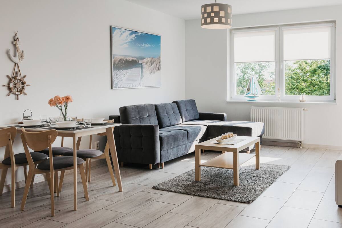 A modern living room with a grey sofa, wooden dining table, and chairs. There's a large window with a view of greenery outside.