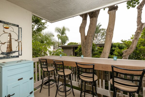 A covered outdoor dining area with a wooden table and four chairs, surrounded by lush greenery and tall trees.