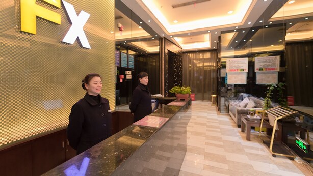 A reception area with a checkered pattern wall and a 'FX' sign. There are two receptionists standing behind a counter. - Fu Yi Fashion Hotel (Chongqing)