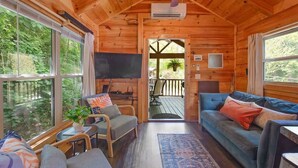 Interior de una cabaña de madera con sofá, sillas, televisor y vistas al exterior.