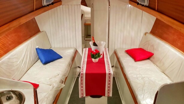 Una cabina stretta e riservata con due divani in pelle bianca, ognuno con un cuscino rosso e blu. Un tavolino con tovaglia rossa contiene una bottiglia e un vaso con fiori.
