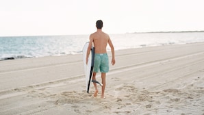 Una persona caminando sobre la arena de una playa, sosteniendo una tabla de surf.