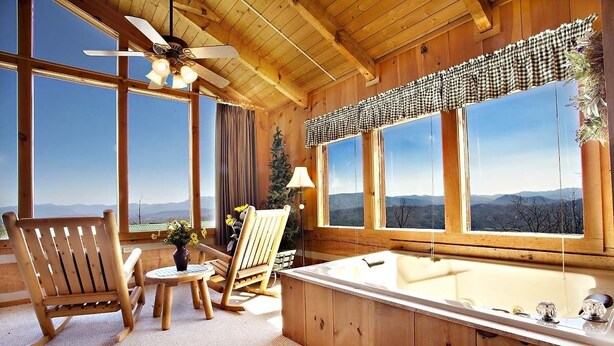 A wooden cabin with a large window, a wooden chair, a small table, and a bathtub. - AWESOME VIEW! 1 bedroom, sleeps 2! (Sevierville)