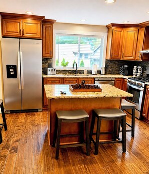 A modern kitchen with wooden cabinets, a stainless steel refrigerator, a built-in island with stools, and a large window.