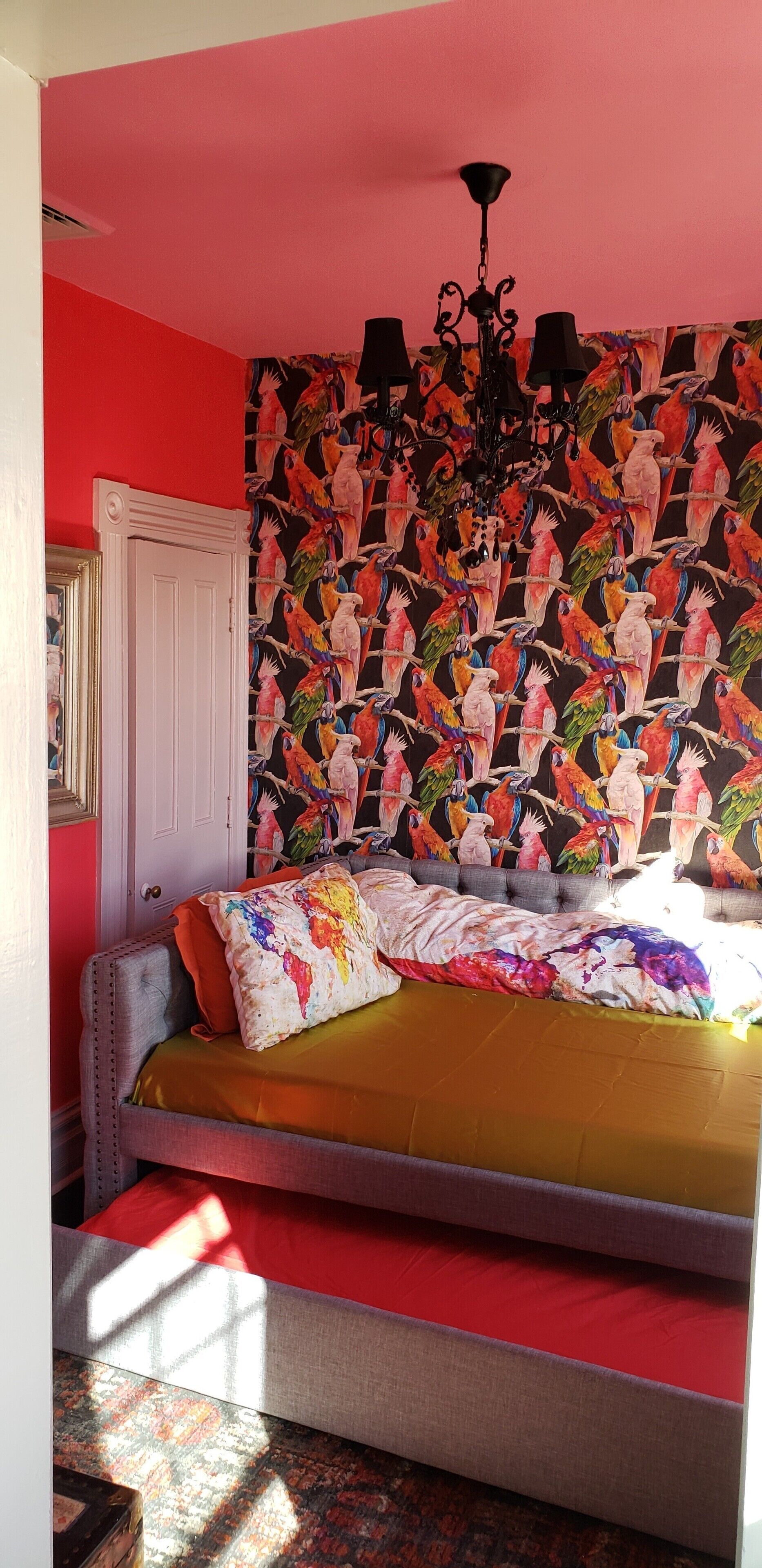 Ein Schlafzimmer mit einer leuchtend roten Wand, einem gemusterten Wandteppich, einem Bett mit vielen Kissen und einem Kronleuchter.