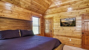 A wooden cabin bedroom with a bed, a window with blinds, a door, and a wall-mounted TV. - Luxury 5BR Cabin w/ Private Pool, Firepit, Theater & Game Room – In Gatlinburg (Gatlinburg)