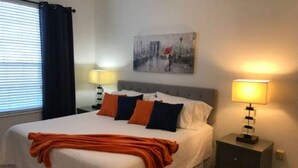 Un lit bien fait, recouvert d’une couverture blanche et orange, avec deux lampes de chevet et un tableau accroché au mur.