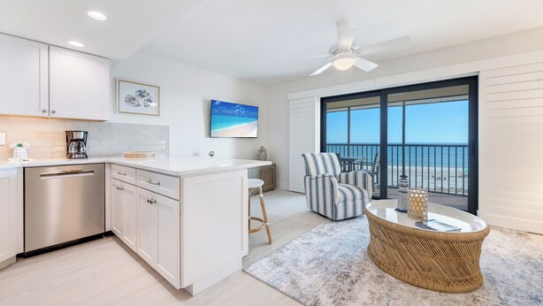 A modern kitchen with white cabinets, a built-in dishwasher, and a dining area with a wicker table and striped chairs. - OCEAN'S REACH 2X4: 1-Bdrm, Top Floor/Gulf Front Unit! All Amenities Open! (Sanibel Island)