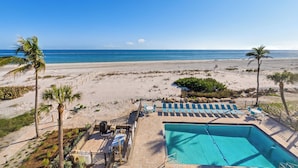 A beachside pool area with lounge chairs and a swimming pool, surrounded by palm trees and a clear view of the ocean. - OCEAN'S REACH 2X4: 1-Bdrm, Top Floor/Gulf Front Unit! All Amenities Open! (Sanibel Island)